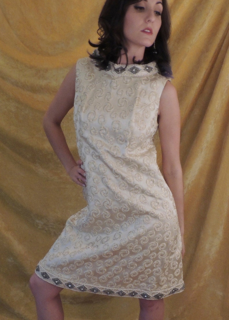 Puede incluir: Un vestido blanco sin mangas con un escote de cuentas y un patr&oacute;n de remolino. El vestido tiene un ribete de cuentas alrededor del dobladillo.