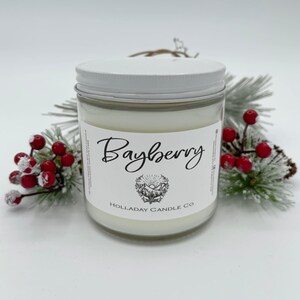 May include: A glass jar candle with a white lid and label that reads "Bayberry." The label features a mountain and tree design. The candle is surrounded by frosted pine branches and red berries.