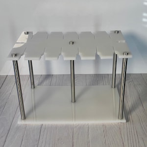 May include: A clear acrylic display stand with a rectangular base and a top shelf. The top shelf has multiple cutouts. The stand has four stainless steel legs and is designed to showcase items. The stand is on a wooden surface.