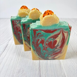 May include: Three bars of handmade soap with a teal, red, and cream swirl design. Each bar is topped with a cream-colored decorative element and a small, orange, lattice-like accent. The soaps are arranged on a white wooden surface.