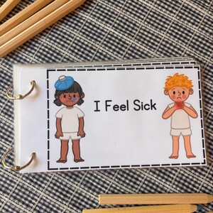 May include: A rectangular laminated card bound with metal rings featuring the text I Feel Sick. The card displays two cartoon figures in white clothing: one with a blue ice pack on their head and a thermometer in their mouth, and another holding their red throat.