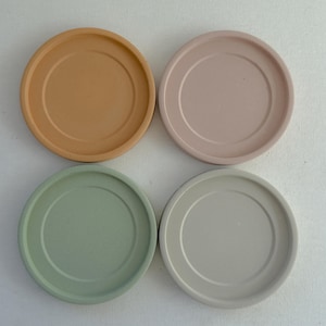 May include: Four round coasters in a top-down view. The coasters are in shades of tan, pink, green, and gray. Each coaster has a raised edge and a recessed center. The coasters are arranged in a square formation.