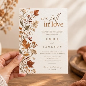 Pode incluir: Um convite de casamento retangular em papel texturizado branco sujo com uma ilustração botânica vertical de folhas de outono e flores secas no lado esquerdo. O texto diz: we fell in love, together with their families you are kindly invited to the wedding of EMMA and JACKSON, Saturday, September 29, 2027, at four o'clock in the afternoon, The Maple Ridge Estate, 123 Autumn Lane, Willow Creek, Colorado, dinner and dancing to follow.