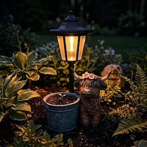 May include: A black solar-powered garden lamp with a warm light illuminates a garden scene. A Highland cow statue with a floral crown and a small metal bucket with a plant are in the foreground. Lush green foliage surrounds the objects.