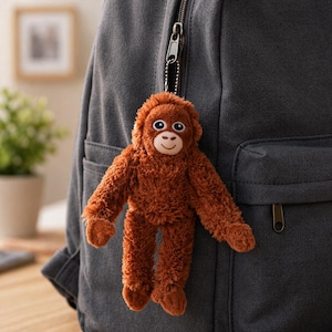 May include: A plush, brown orangutan keychain with a smiling face and outstretched arms hangs from a gray backpack. The keychain features a metal chain and a zipper pull.