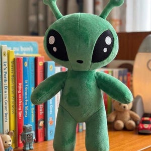Puede incluir: Un juguete de peluche verde alienígena con grandes ojos negros y antenas. El peluche está de pie sobre una estantería de madera frente a una estantería. El alienígena tiene un diseño cosido y es un juguete para niños.