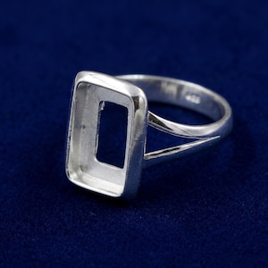 May include: A silver ring with a rectangular setting, designed to hold a gemstone. The ring has a split shank and a polished finish. The setting is open, allowing light to pass through the stone.