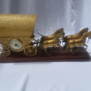 May include: A gold-toned covered wagon clock featuring a textured, arched top and four spoked wheels. The wagon is pulled by four gold-toned horse figures attached to a dark brown rectangular wooden base. The clock face is circular with black numerals.