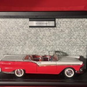 May include: A red and white classic convertible car model with chrome detailing, presented in a black display case against a brick wall background. The model has white-walled tyres and a detailed interior. A small plaque above the car reads "Ford Fairlane".