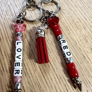 May include: Keychains with silver hardware, featuring beaded accents and charms. One keychain spells "LOVER" in white letter beads, another says "RED", and a third has a red tassel. The keychains have heart-shaped and red bead details.