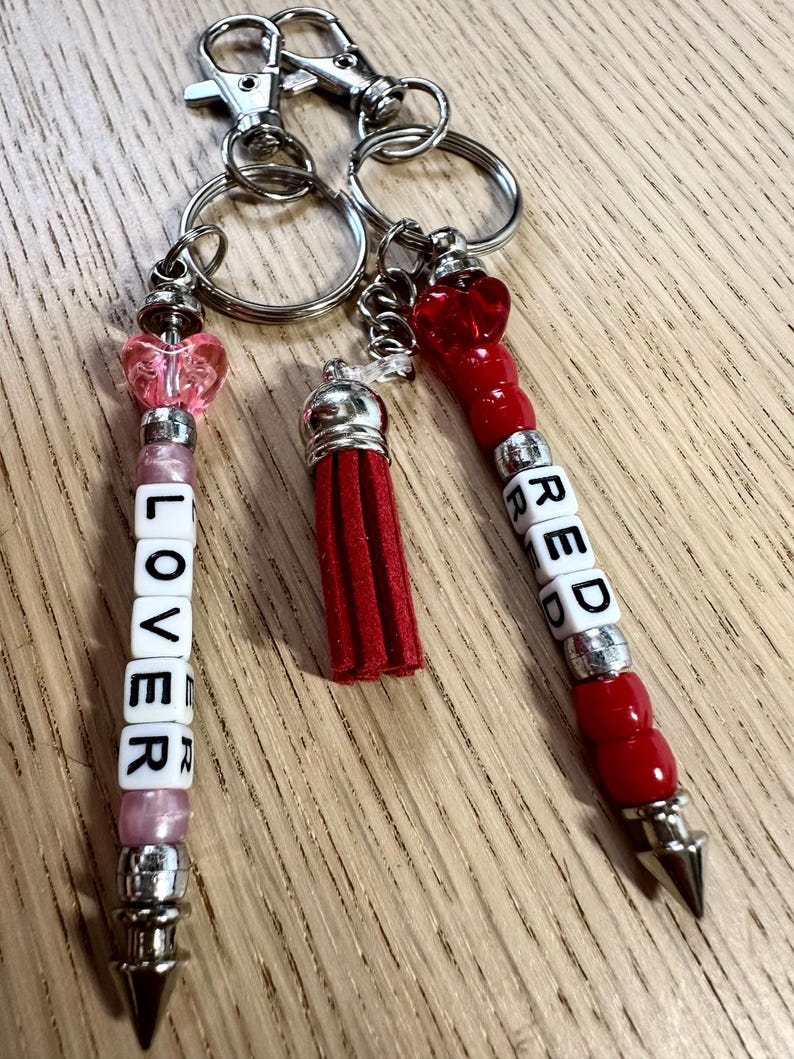 May include: Keychains with silver hardware, featuring beaded accents and charms. One keychain spells "LOVER" in white letter beads, another says "RED", and a third has a red tassel. The keychains have heart-shaped and red bead details.