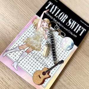 May include: A keychain with a guitar charm, a disco ball charm, and a charm that says "Reputation" is displayed on a word search puzzle book. The book cover features an illustration of a blonde woman in a gold dress. The book is titled "Taylor Swift Word Search and Color."