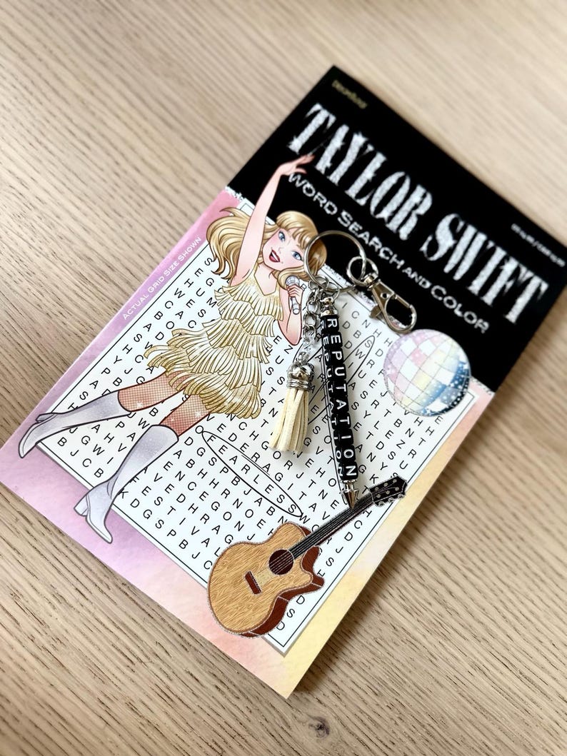 May include: A keychain with a guitar charm, a disco ball charm, and a charm that says "Reputation" is displayed on a word search puzzle book. The book cover features an illustration of a blonde woman in a gold dress. The book is titled "Taylor Swift Word Search and Color."