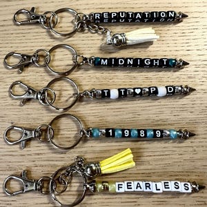 May include: Keychains with silver-tone hardware and beaded designs. The keychains feature the words "REPUTATION", "MIDNIGHT", "TTPD", "1989", and "FEARLESS" spelled out with letter beads. Some keychains have decorative tassels.
