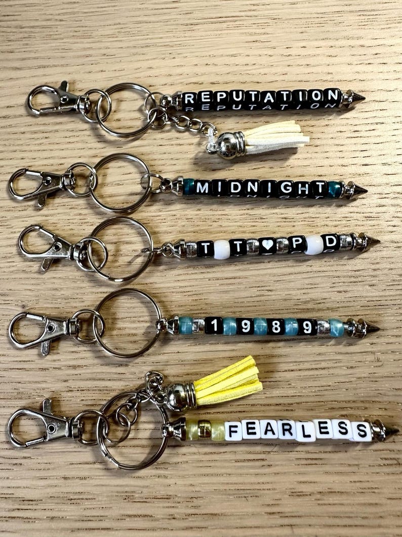 May include: Keychains with silver-tone hardware and beaded designs. The keychains feature the words "REPUTATION", "MIDNIGHT", "TTPD", "1989", and "FEARLESS" spelled out with letter beads. Some keychains have decorative tassels.