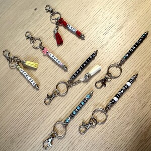May include: A collection of keychains with beaded pen-shaped charms. Each keychain features a different word spelled out in white letter beads, including "FEARLESS", "LOVER", "REPUTATION", "MIDNIGHT", "1989", and "RED". Some have tassels.
