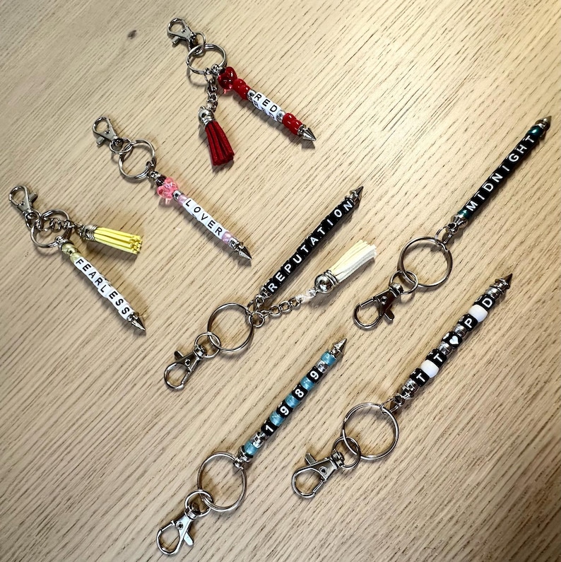 May include: A collection of keychains with beaded pen-shaped charms. Each keychain features a different word spelled out in white letter beads, including "FEARLESS", "LOVER", "REPUTATION", "MIDNIGHT", "1989", and "RED". Some have tassels.