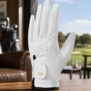 May include: A white golf glove featuring perforated detailing along the fingers and back of the hand. The wrist closure includes a hook-and-loop fastener tab with the gold-colored letters HR printed on the front, finished with a black trim edge.