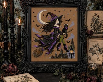 Puede incluir: Un bordado en punto de cruz enmarcado que representa a una bruja de pelo oscuro con vestido y sombrero negros con detalles morados, montada en una escoba con un gato negro. La escena incluye una luna creciente, estrellas, murciélagos y la silueta de un castillo.