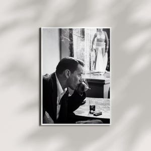 May include: A black and white photograph showing a person in a dark suit jacket and light shirt sitting at a table, holding a cigarette. On the table sits a small glass and a rectangular pack. In the background, a figure stands on a stage.