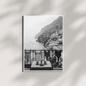 May include: A black and white wall art print depicting a coastal balcony scene. Four one-piece swimsuits hang on a railing above a small table set with two cups and a plate. In the background, a hillside town overlooks a bay with small boats.