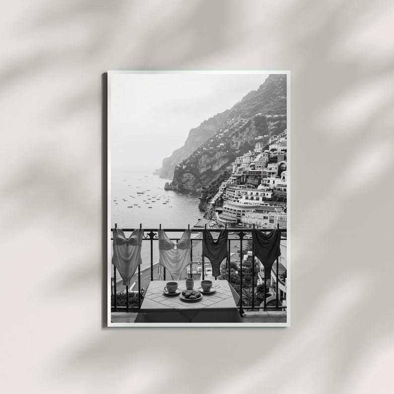 May include: A black and white wall art print depicting a coastal balcony scene. Four one-piece swimsuits hang on a railing above a small table set with two cups and a plate. In the background, a hillside town overlooks a bay with small boats.