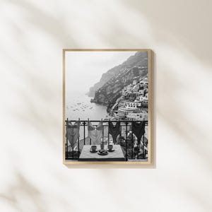 May include: A black and white framed wall art print depicting a coastal scene with four swimsuits hanging on a balcony railing overlooking a hillside town and the sea. A small table with two cups and a plate sits on the balcony in the foreground.