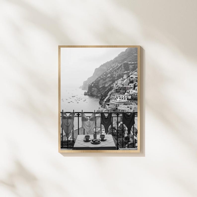 May include: A black and white framed wall art print depicting a coastal scene with four swimsuits hanging on a balcony railing overlooking a hillside town and the sea. A small table with two cups and a plate sits on the balcony in the foreground.
