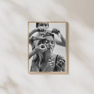 May include: A black and white photographic print featuring a person with long hair being attended to by multiple hands. One hand holds a cotton swab near the eye, while another hand holds a lit cigarette to the person's lips in a frame.