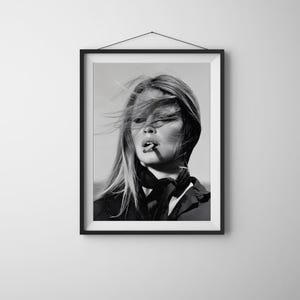 May include: A black and white framed wall art print featuring a portrait of a person with long hair blowing in the wind, wearing a dark collared shirt with a neck scarf, and holding a cigarette between their lips, presented in a simple frame.