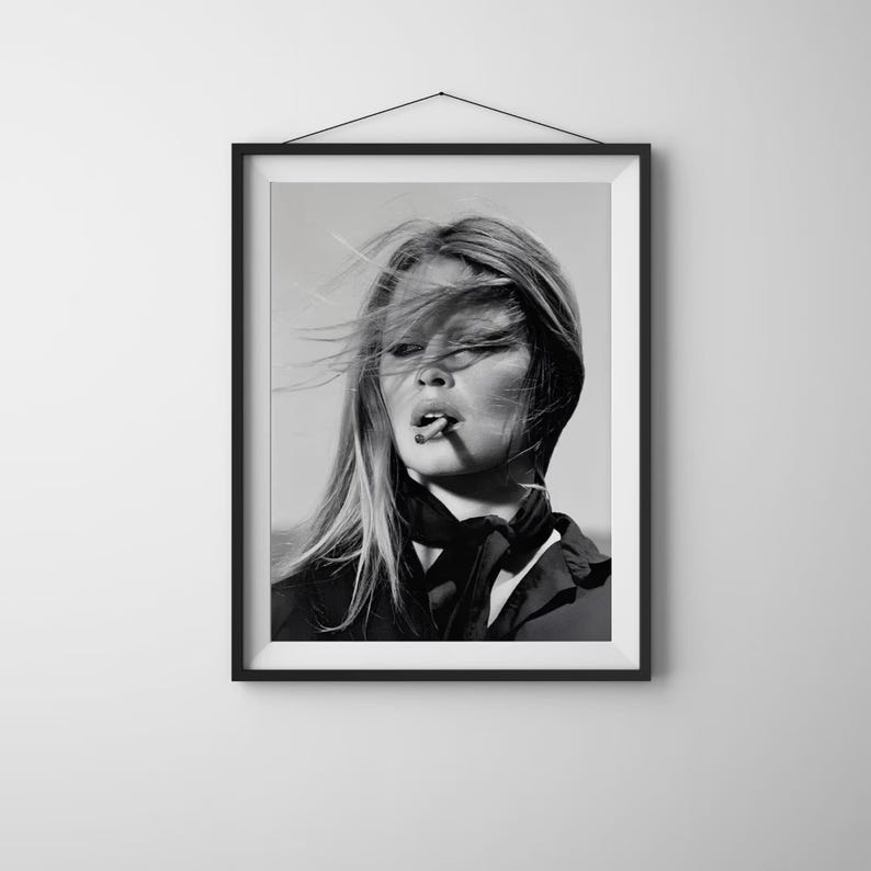 May include: A black and white framed wall art print featuring a portrait of a person with long hair blowing in the wind, wearing a dark collared shirt with a neck scarf, and holding a cigarette between their lips, presented in a simple frame.