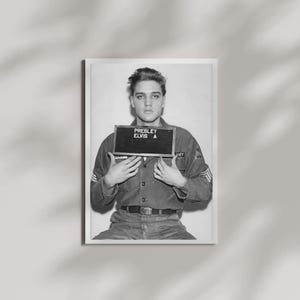 May include: A black and white photograph of a person in a military uniform holding a sign that reads PRESLEY ELVIS A. The individual has dark hair and is wearing a button-down shirt with a belt, captured in a front-facing portrait style.