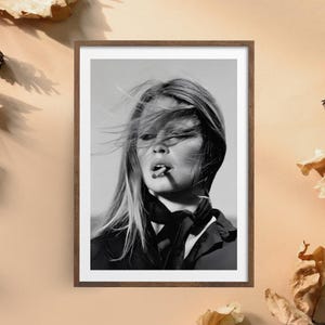 May include: A black and white framed portrait photograph of a person with long hair blowing in the wind, wearing a dark collared shirt with a necktie. The person has a cigarette held between their lips and is looking slightly away from the camera.