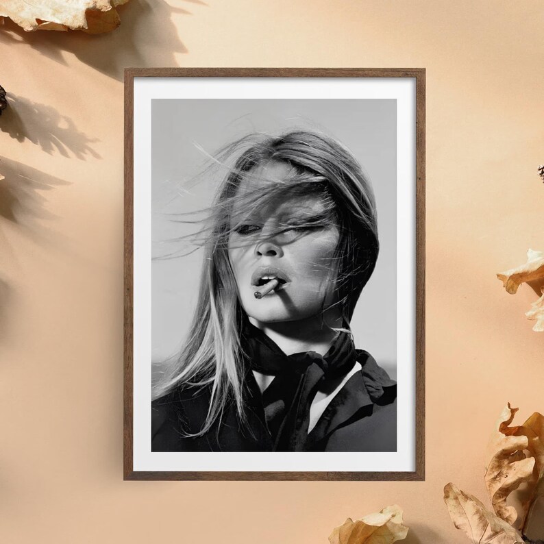 May include: A black and white framed portrait photograph of a person with long hair blowing in the wind, wearing a dark collared shirt with a necktie. The person has a cigarette held between their lips and is looking slightly away from the camera.