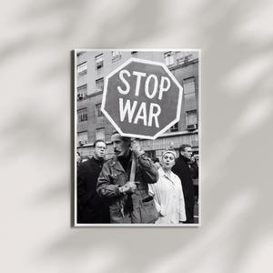 Op de afbeelding: Een zwart-witfoto van een groep mensen die deelneemt aan een protest. Een centraal figuur houdt een groot, achthoekig bord vast met de tekst STOP WAR. De personen dragen jassen en staan voor een gebouw.