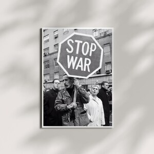 Op de afbeelding: Een zwart-witfoto van een groep mensen die deelnemen aan een protest. Een persoon op de voorgrond houdt een groot achthoekig bord vast met de tekst STOP WAR in witte hoofdletters, met een stadsgebouw op de achtergrond.