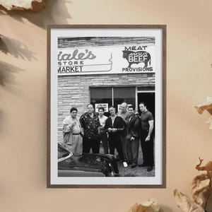 May include: A black and white framed print depicting a group of six men standing in front of a brick storefront labeled 'Meat Angus Beef Provisions'. The men are dressed in casual attire, and the front of a dark car is visible in the foreground.