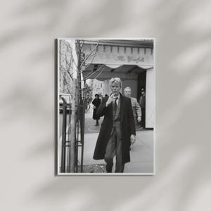Op de afbeelding: Een zwart-witfoto van David Bowie die over een stadsstoep loopt voor hotel The Carlyle. Hij draagt een lange donkere jas, een vest met patroon en een overhemd met kraag. Andere figuren zijn zichtbaar op de achtergrond.