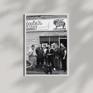 May include: A black and white photograph of eight individuals standing in front of a brick storefront labeled 'Satriale's Pork Store' and 'MEAT ANGUS BEEF PROVISIONS'. A portion of a dark car is visible in the foreground, and the group is posed together.