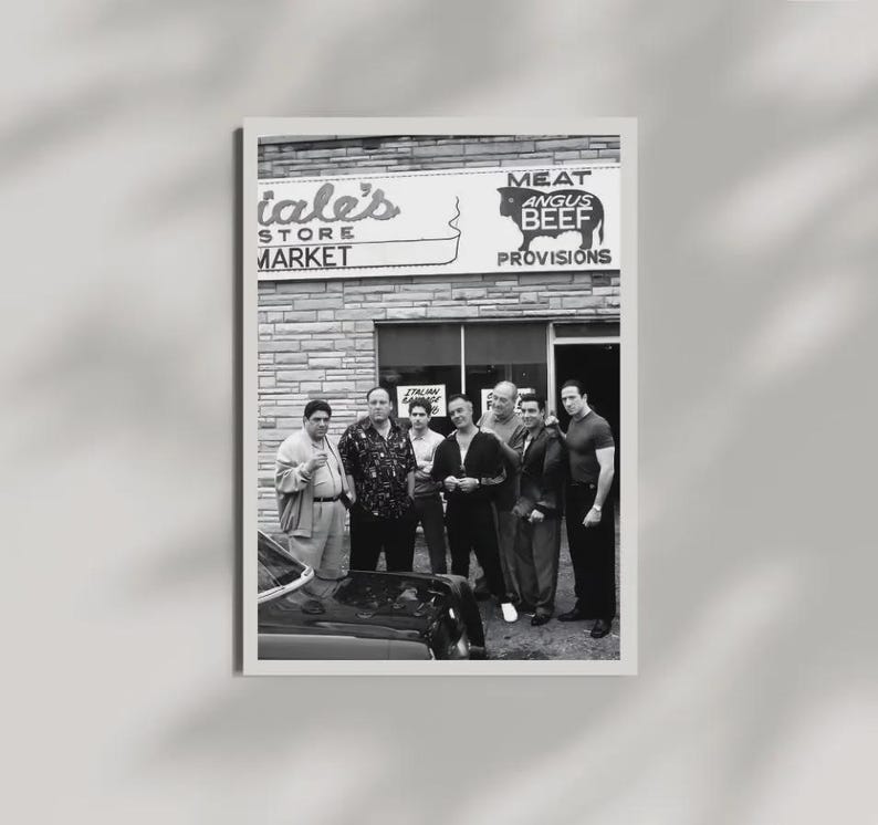 May include: A black and white photograph of eight individuals standing in front of a brick storefront labeled 'Satriale's Pork Store' and 'MEAT ANGUS BEEF PROVISIONS'. A portion of a dark car is visible in the foreground, and the group is posed together.