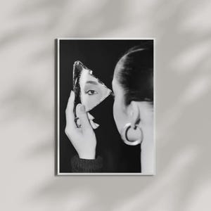 May include: A black and white photographic print showing a side profile of a head with a large hoop earring. A hand holds a triangular shard of a mirror, reflecting a single eye, against a dark background. The composition is high contrast.