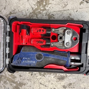 May include: A black and clear plastic case with a red interior contains plumbing tools. The tools include a blue and black Geberit press tool, metal crimping tools, and red accessories. The case is open, revealing the tools.