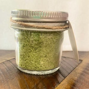 May include: A small, clear glass jar filled with green granules, likely bath salts or a similar product. The jar has a silver metal lid, a twine accent, and a small, blank tag. The jar sits on a wooden surface.