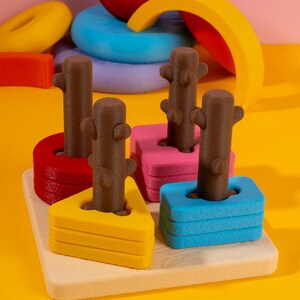 May include: A colorful wooden stacking toy with a light wood base. The toy features brown tree trunk shapes that fit into red, pink, yellow, and blue geometric shapes. Other colorful wooden shapes are in the background.