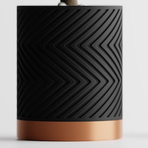 May include: A cylindrical object with a black, textured surface featuring a chevron pattern. The base is a copper color. The object is set against a white background.