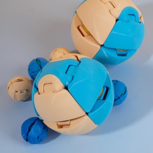 Könnte beinhalten: Mehrere kugelförmige Puzzles in Blau- und Beigetönen liegen auf einer hellblauen Oberfläche. Jedes Puzzle besteht aus ineinandergreifenden, gebogenen Segmenten. Das Bild zeigt Puzzles in verschiedenen Größen, die 3D-gedruckt zu sein scheinen. Ein spielerisches Objekt.