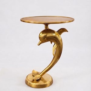 Può includere: Un tavolino in metallo color oro con piano circolare sostenuto da una base scultorea a forma di delfino che salta. Il delfino è posizionato verticalmente, con la coda che poggia su una base rotonda e piatta e la testa che sostiene il piano.