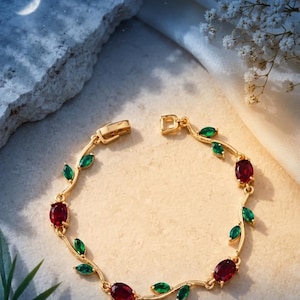 May include: A gold-toned bracelet featuring a vine-like design with alternating oval-shaped red stones and marquise-cut green stones. The metal links connect the floral-inspired elements, and the piece includes a secure box clasp closure.