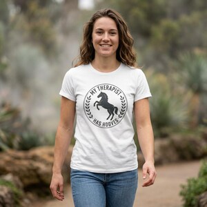Op de afbeelding: Wit T-shirt met de tekst "MY THERAPIST HAS HOOVES" in een cirkelvormig ontwerp met een zwart paard. Het shirt wordt gedragen door een persoon in een blauwe spijkerbroek, die buiten loopt.