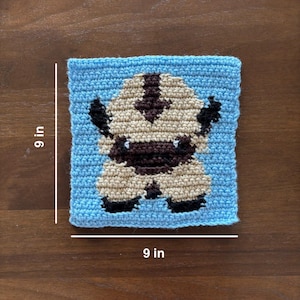 May include: A square crochet piece measuring 9 inches by 9 inches, featuring a light blue background with a central graphic of a beige, brown, and black creature with a dark arrow marking on its head, crafted in a textured yarn stitch pattern.
